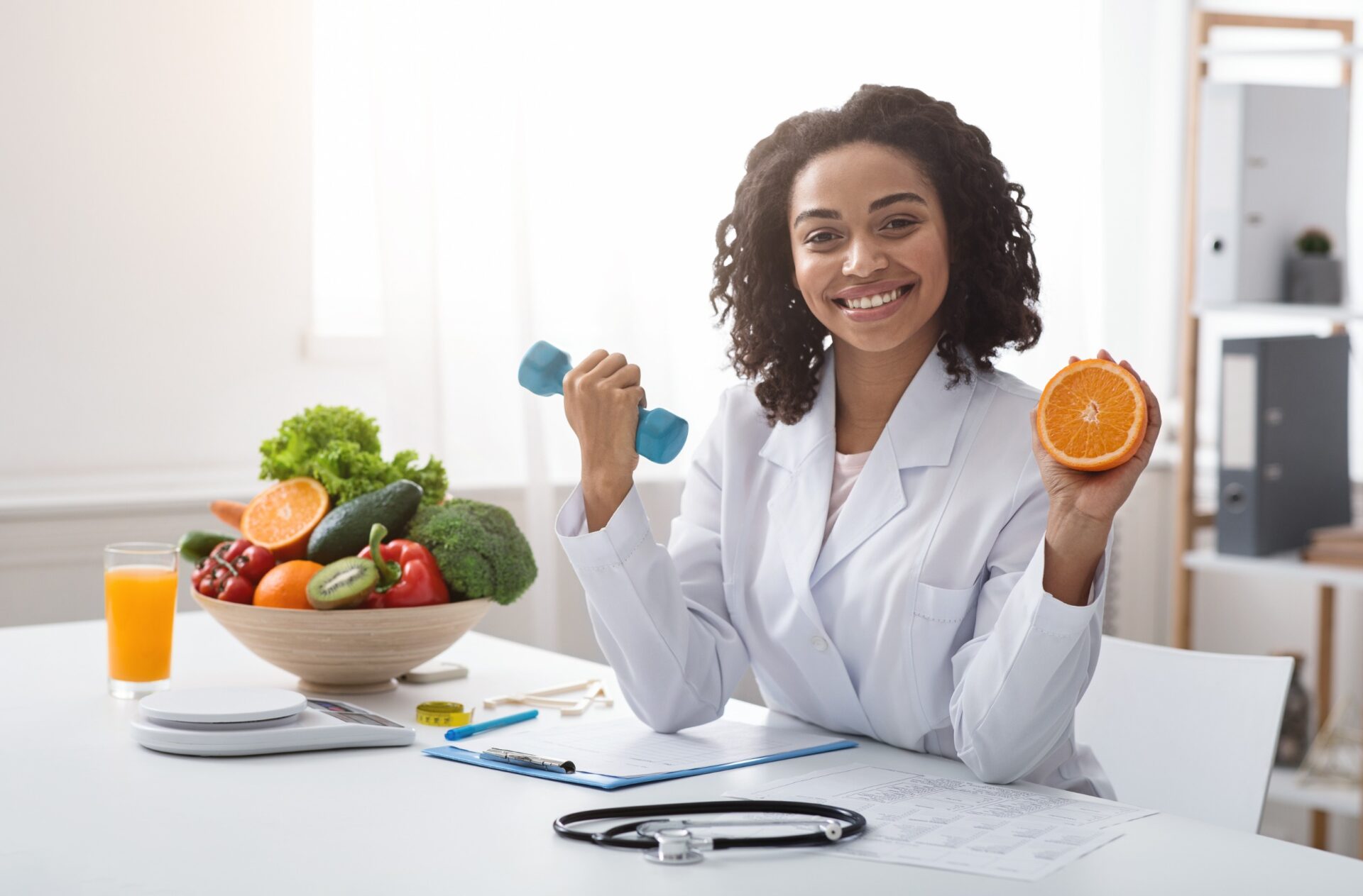 Sport and vitamins for immunity. Happy black woman doctor holding orange and barbell in clinic, posing at her workplace, free space